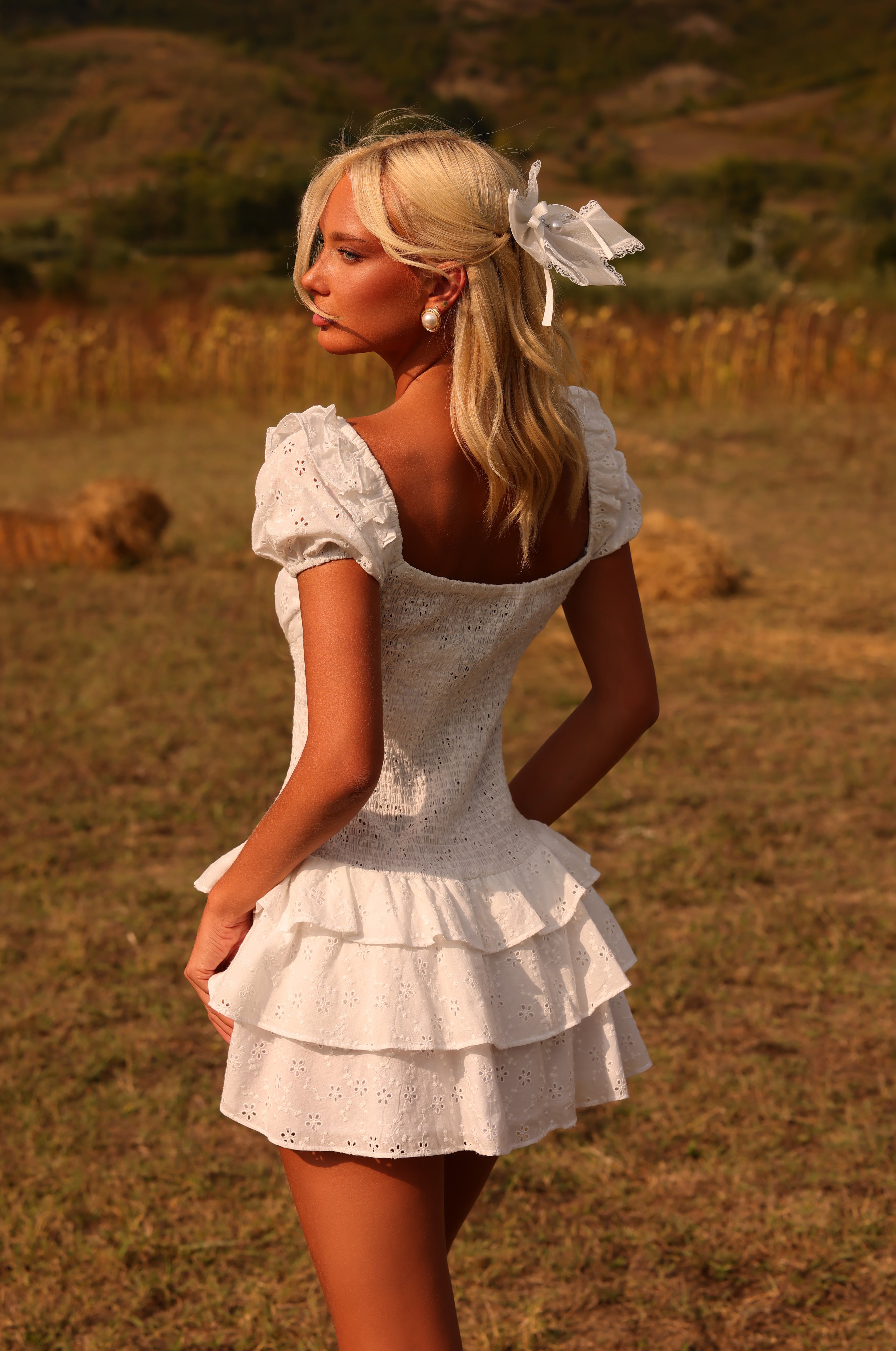 A woman with blonde hair, wearing the Ivory White Cotton Mini Graduation Dress: Macie and a hair ribbon, stands outdoors in a field with hay bales and hills in the background.